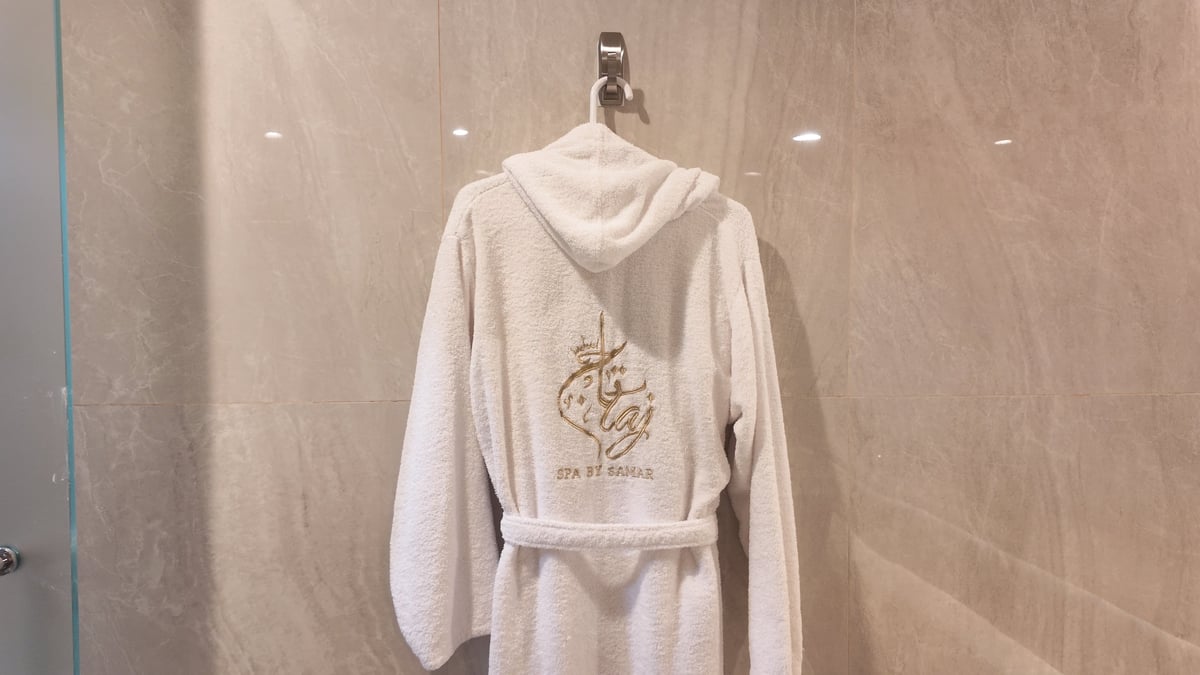 Cream-colored terrycloth robe with hood hanging on bathroom wall hook, showing embroidered design on back